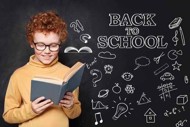 Happy child reading a book in classroom on chalkboard background Back to school concept