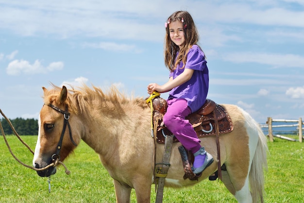 幸せな子供は、背景に青い空と美しい自然を持つ農場の動物の茶色のポニーに乗る