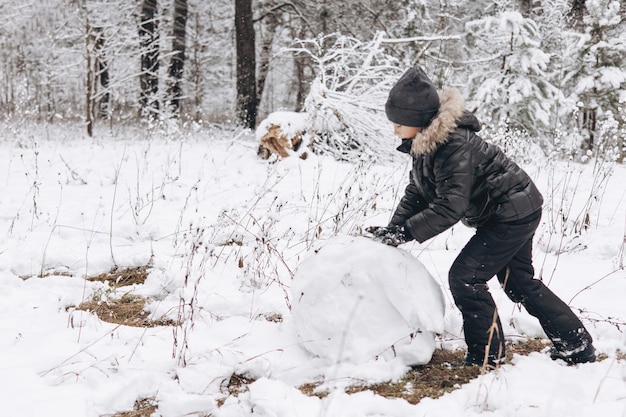 Happy child rolling big snowball for snowman in snowy winter forest. Teenager boy playing and having fun on walking in frosty day. Wintertime activity outdoors.