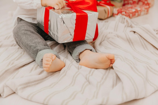 A happy child in a Santa hat sits on the floor near the Christmas tree and holds a box with a gift in his hands. bare heels close up