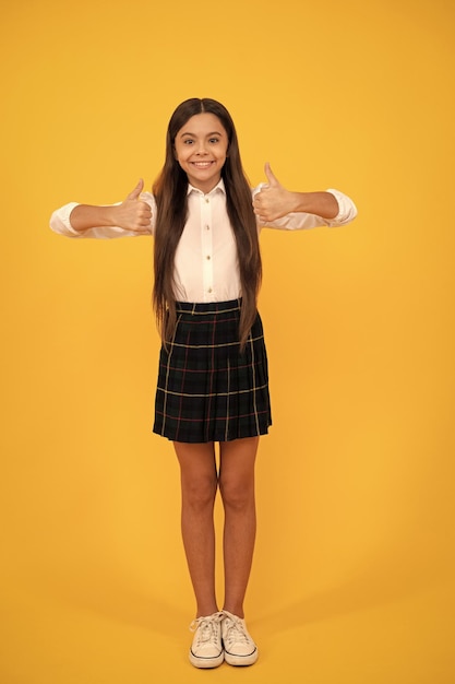 Happy child in school uniform full length showing thumbs up on yellow background positive emotions