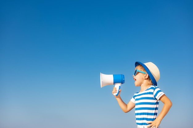 Happy child shouting through loudspeaker outdoor against blue sky background. Portrait of smiling kid on summer vacation. Freedom and travel concept