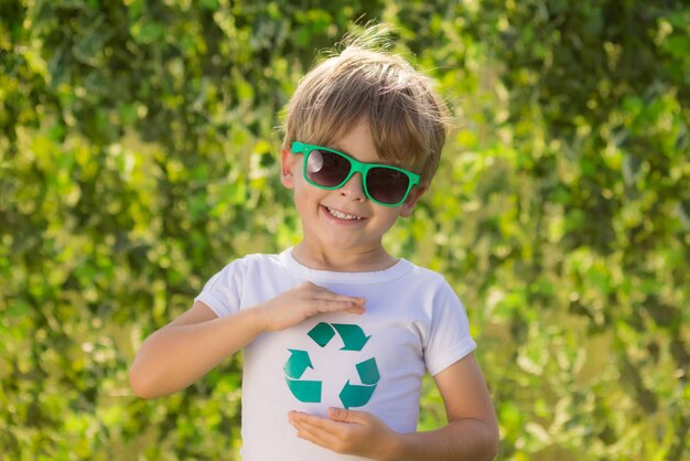 Счастливый ребенок показывает знак переработки. Портрет ребенка на открытом воздухе. Мальчик на размытом зеленом весеннем фоне. День земли и концепция экологии
