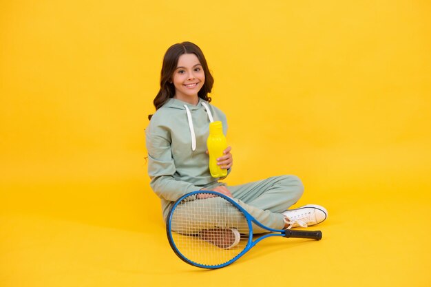 Happy child sit in sportswear with tennis racquet and water bottle on yellow background, water balance.