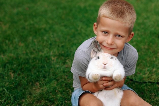 Happy child sits on the green grass and holds a rabbit in his arms.