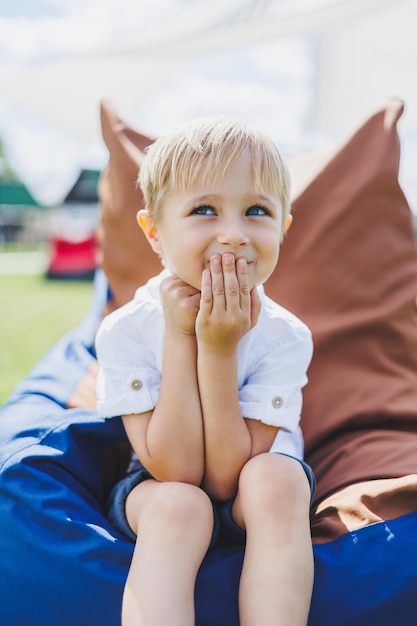 Счастливый ребенок сидит в кресле-мешке в летнем парке Маленький мальчик в белой футболке веселится на улице Детство и счастье