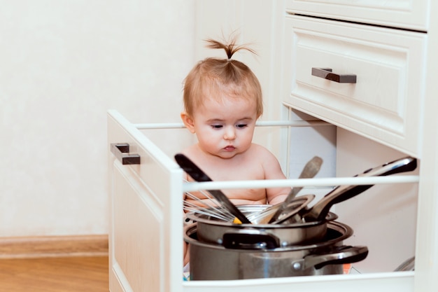 Happy child sitting in the kitchen drawer with pots and laughing. Portrait of a toddler in a white kitchen.