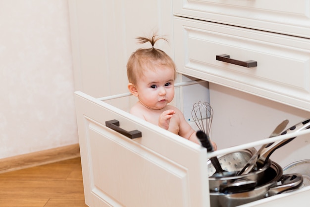 鍋を持って台所の引き出しに座って笑っている幸せな子供。白いキッチンで幼児の肖像画。