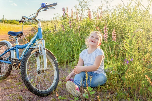 自転車のそばに座って幸せな子。日当たりの良い夏の公園で自転車を持つ少女