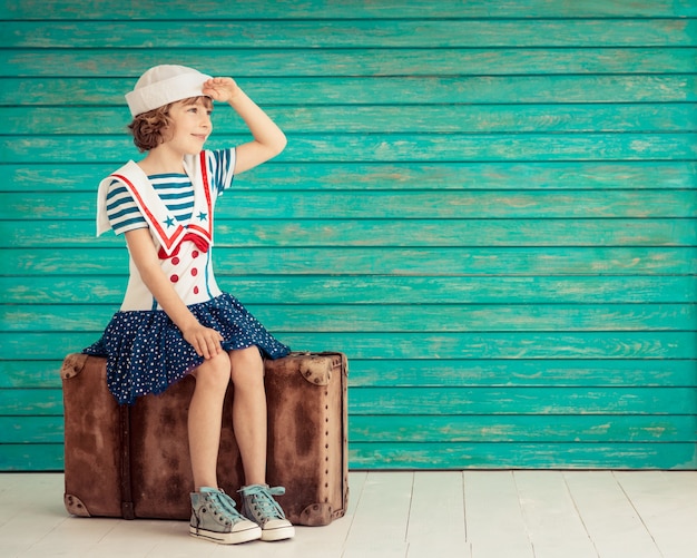 happy child sitting on an old suitcase dressed like a sailor looking ahead on blue wooden background