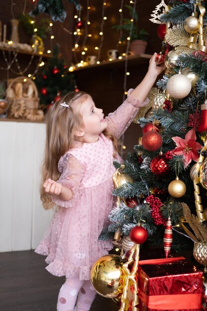 Happy child smiling blond girl in beautiful dress decorating christmas tree