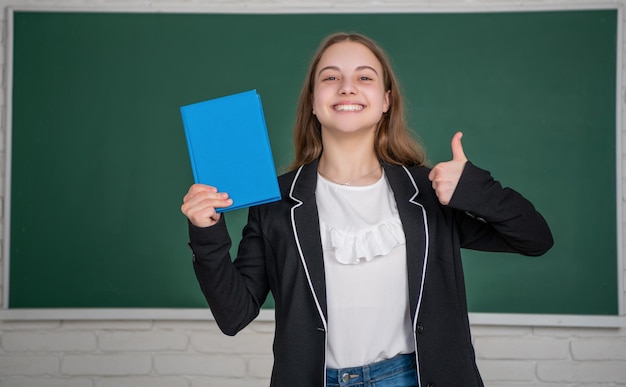Happy child standing on blackboard background with workbook at school thumb up