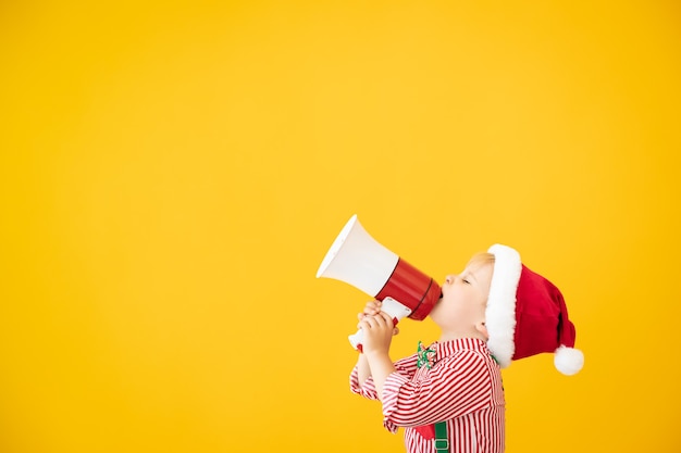 Happy child wearing santa claus costume speaking by megaphone. portrait of funny kid against yellow background