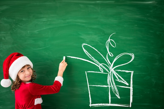 Happy child wearing Santa Claus draws on green blackboard