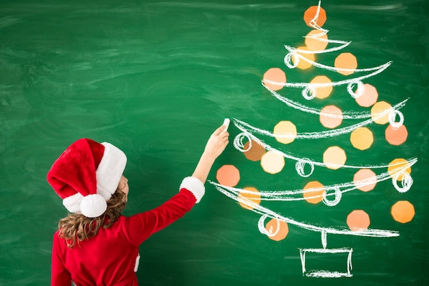 Happy child wearing Santa Claus draws on green blackboard