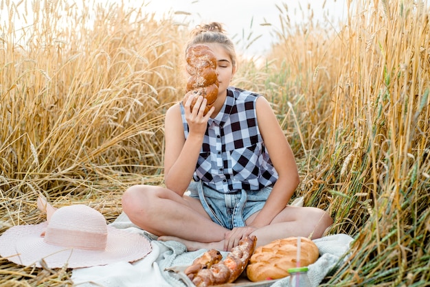 Photo happy child with bread in yellow autumn wheat field. a field with mature ears. a girl sits on a bedspread, fresh fruits and berries, bread and rolls in a basket. outdoors village picnic.