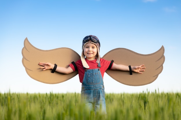 Happy child with cardboard wings playing outdoor in green field Portrait of kid against summer blue sky