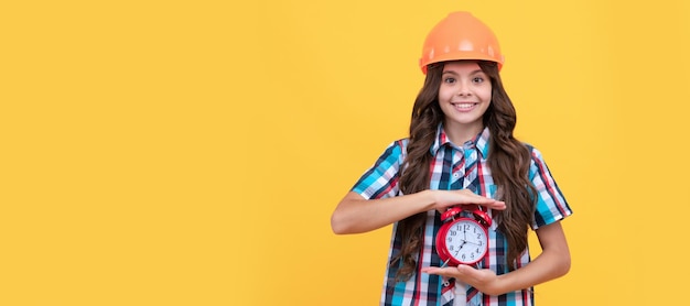 Happy child with curly hair in construction helmet showing alarm clock timing Child builder helmet