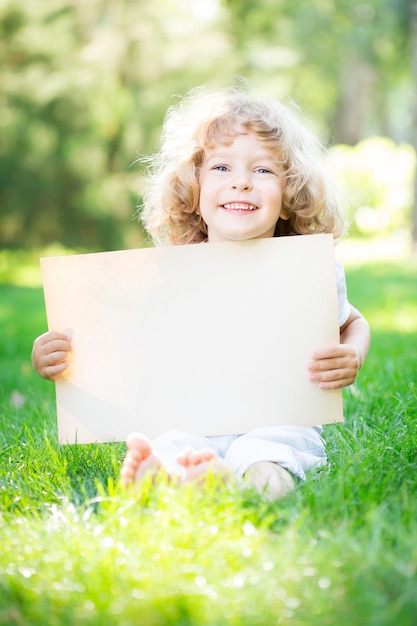春の公園の緑の芝生の上に座っている紙の空白の幸せな子