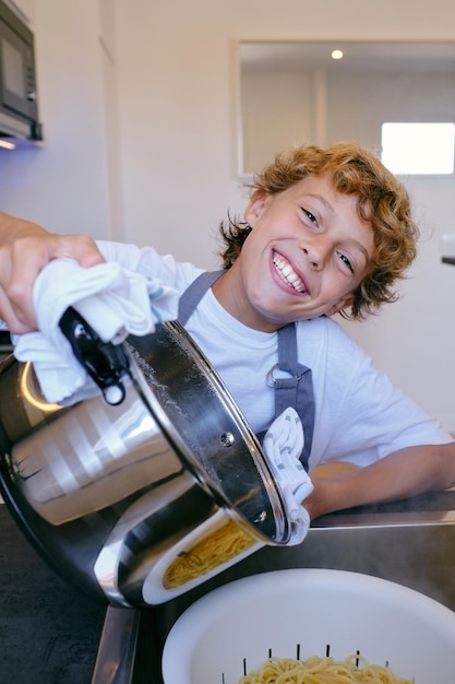 Happy child with saucepan above strainer with cooked pasta in sink looking at camera at home