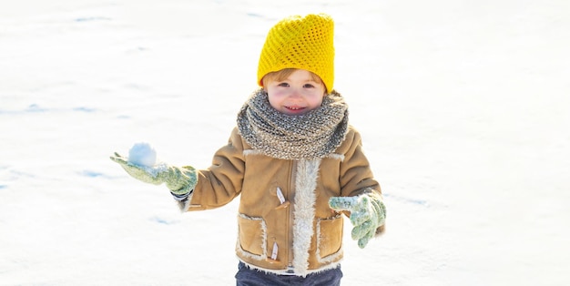 Happy child with snowball on snow background cute kid boy in winter clothes hat and scarf close up c