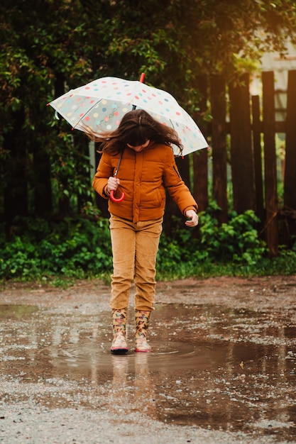 Photo happy child with an umbrella and rubber boots jump in puddle on a walk