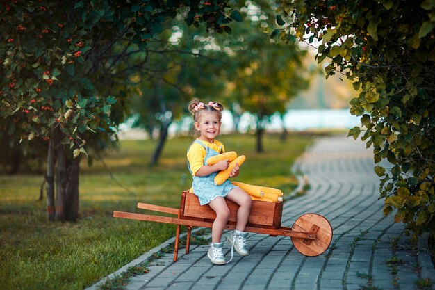 Счастливый ребенок с початками желтой мозоли в тачке. Красивая девочка с початками кукурузы. Ребенок счастливый Осенний урожай в деревянной тачке.