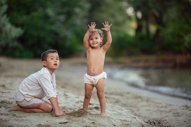 Счастливое детство, смешно. Дети брат и сестра играют на песке на берегу реки возле зеленых деревьев летом на отдыхе.