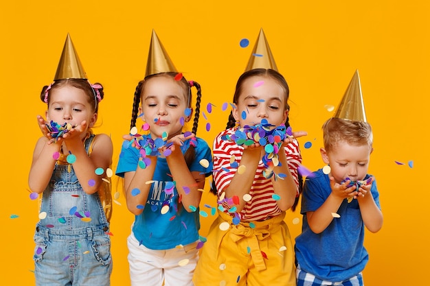 Foto i bambini felici in vacanza si divertono e saltano in coriandoli multicolori su sfondo giallo