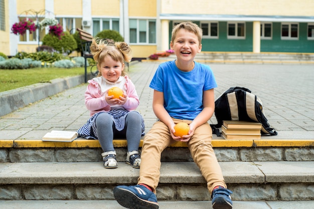 Счастливые дети, играющие в школьном дворе в дневное время. школьный завтрак, фрукты и сок. стек учебников, книг. счастливые дружелюбные сестра и брат