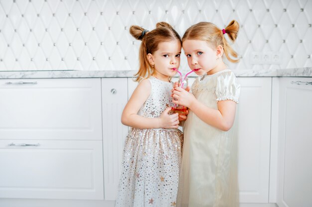 happy children in a white kitchen drink apple juice from a glass bottle through the straws