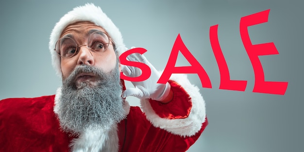 Happy Christmas Santa Claus on grey studio background. Caucasian male model in traditional holiday's costume. Concept of holidays, new year's, winter mood, gifts. Christmas sale. Copyspace.