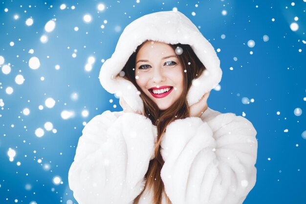 Foto buon natale e vacanze invernali ritratto di giovane donna in pelliccia bianca con cappuccio neve su sfondo blu moda e stile di vita