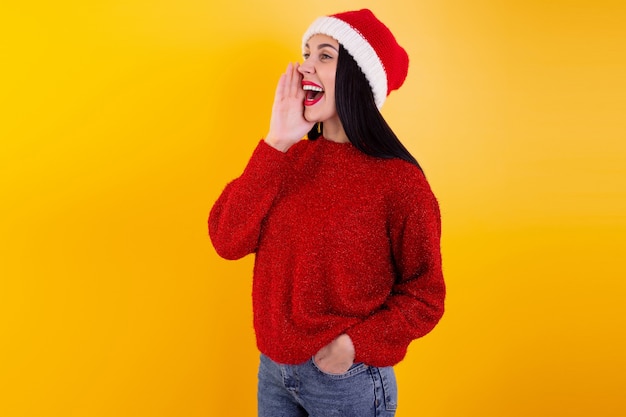 Happy christmas woman excited say hello isolated on yellow background wearing red santa hat, shouts discounts, holding his hand around his mouth. christmas discount content.