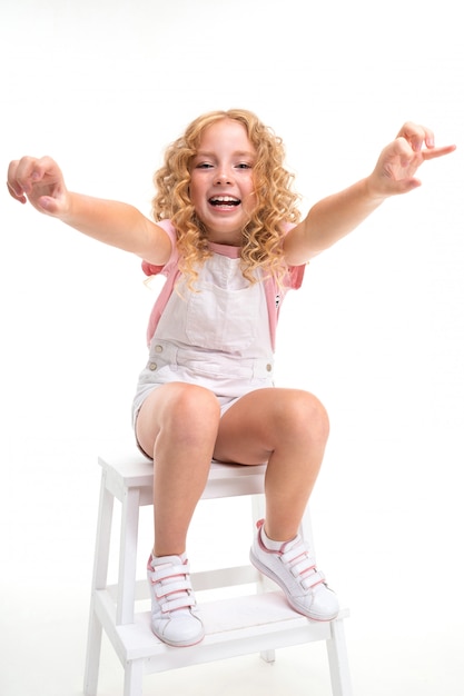 Happy cild with fair red curly hair sits on a chair and smile isolated on white surface