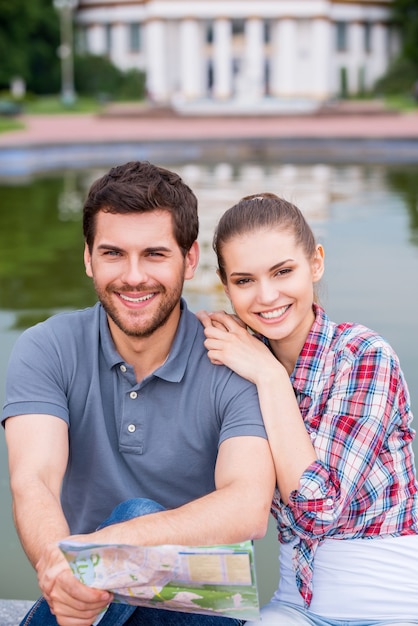 Happy city travelers. Top view of happy young tourist couple sitting near beautiful building and looking at camera while man holding map