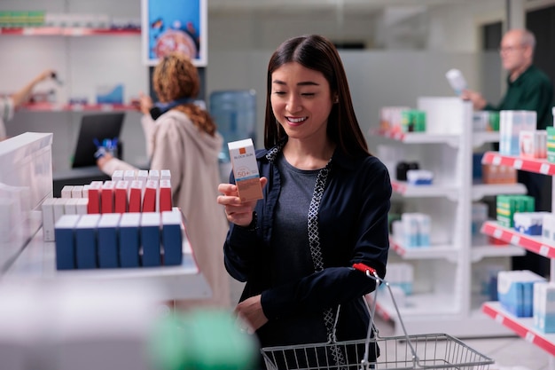 Happy client holding SPF face cream reading package prescription before choosing to buy it. Smiling customer holding basket shopping for pharmaceutical vitamin in pharmacy shop