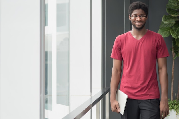 행복한 대학생이 학교로 돌아갑니다. 행복한 미소 짓는 괴짜 남자, 대학 캠퍼스에서 노트북 컴퓨터를 가진 아프리카 대학생의 초상화, 다시 학교 개념으로; 아프리카 젊은 성인 남자 모델