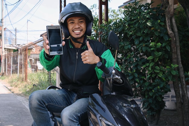 Happy commercial motorcycle taxi driver holding smart phone with blank screen for mockup and showing