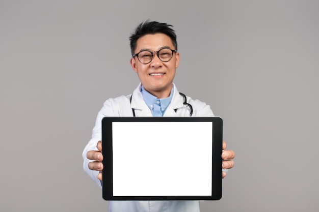 흰색 코트와 안경을 쓴 행복한 자신감 있는 성인 일본 의사는 빈 화면이 있는 태블릿을 보여줍니다.
