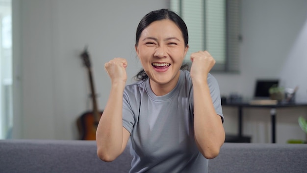 Happy confident asian woman sitting on sofa looking at camera\
showing her fist make a winning gesture