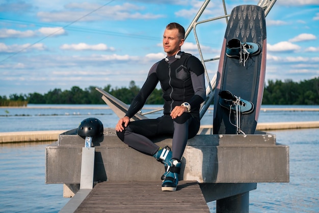 Happy confident man in wetsuit sitting on pier with wakeboard