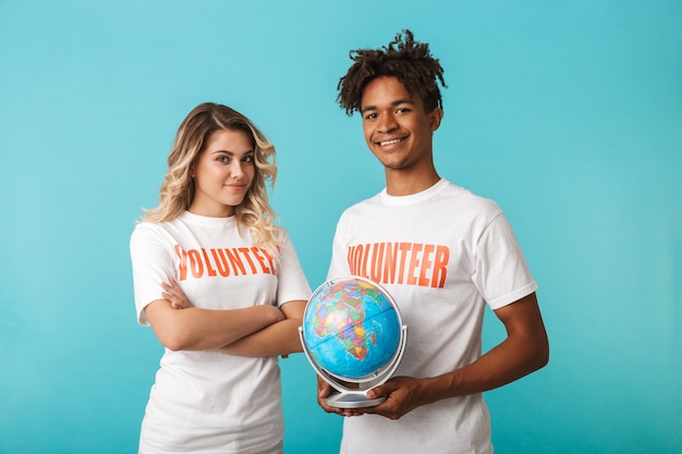 Happy confident multiethnic couple wearing volunteers t-shirt standing isolated over blue wall, holding a globe