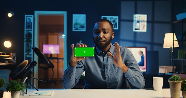 Счастливый создатель контента с Chroma Key продуктом, спонсируемым технологической компанией для съемок обзора разбокса афроамериканский влиятельный человек, показывающий макет экрана телефона рекомендация для зрителей