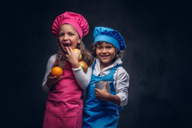 Piccola coppia di cuoco felice. un ragazzino con capelli ricci marroni vestito con un'uniforme da cuoco blu e una bella studentessa vestita con un'uniforme da cuoco rosa, posa con pentole e frutta in uno studio. isolato