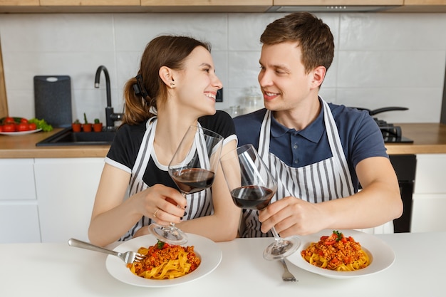 Happy couple in aprons eating bolognese pasta with wine at modern kitchen Romantic dinner at home