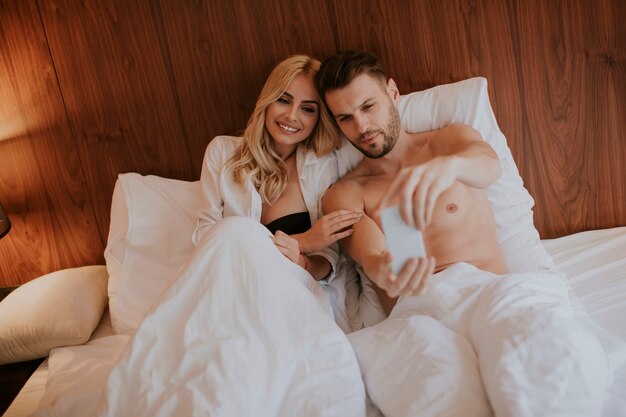 Happy couple in bed at home and use mobile phone