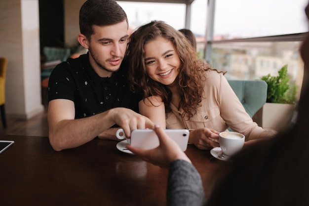 Happy couple in cafe look into smartphone and smile Woman show to man and woman something in phone Man put finger on screen and smile Woman laughing