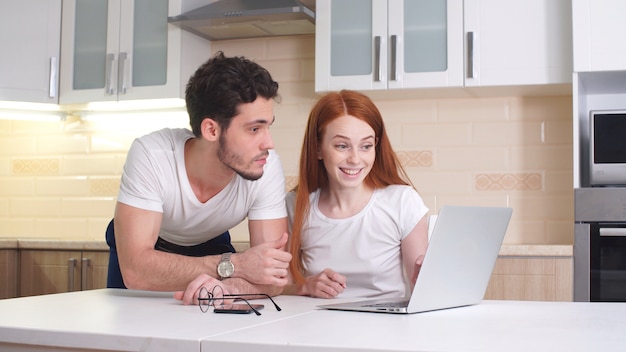 Счастливая пара выбирает, куда поехать в отпуск, смотрит на ноутбук, сидит дома на кухне