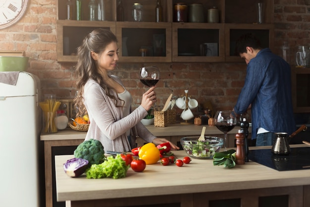 행복한 커플은 집에 있는 로프트 주방에서 건강식을 함께 요리합니다. 와인을 마시는 여자. 야채 샐러드 준비.